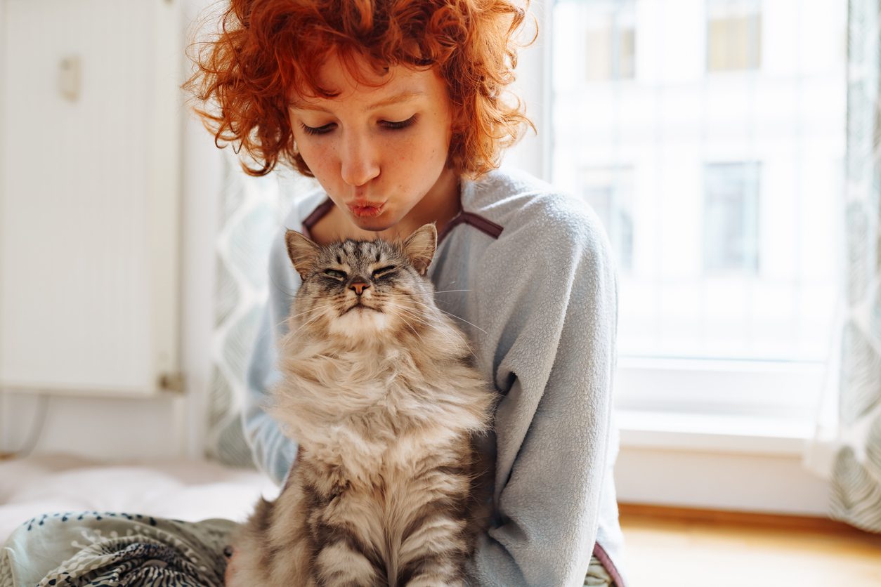 red-haired girl teenager hugs kisses a purring Maine Coon cat, sitting on floor near window at home